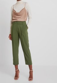 Camiseta de satén marrón sobre un cuello de tortuga crema, combinada con pantalones de talle alto verdes y tacones de tiras nude.