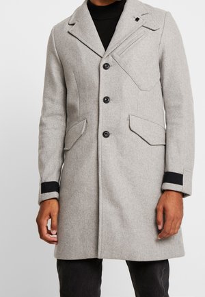 Cappotto grigio in lana con un design su misura, caratterizzato da un colletto a punta, tre bottoni, tasche laterali e dettagli neri sui polsini.