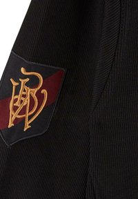 Un vêtement tricoté noir avec un patch rectangulaire ornée d'un design doré brodé et une bande bordeaux. Surface texturée avec des détails côtelés.