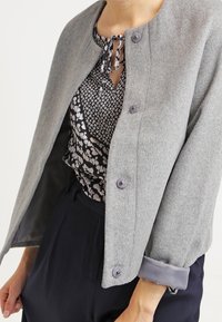 Chaqueta de mezcla de lana gris con cuello redondeado, cuatro botones y mangas abullonadas. Llevada sobre una blusa estampada con detalle de lazo.
