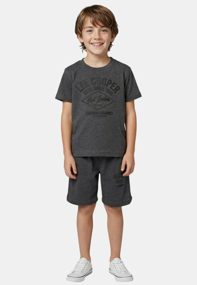 Garçon souriant debout, portant un t-shirt gris foncé Lee Cooper et un short assorti avec des baskets blanches, sur un fond clair uni.
