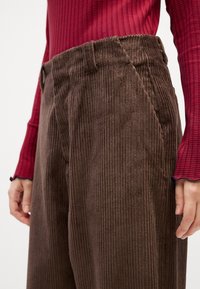 Brown corduroy trousers featuring vertical ridges, a high waist, and side pockets. Paired with a fitted red and navy striped top.