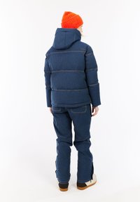 Persona che indossa una giacca imbottita blu navy con cappuccio, pantaloni coordinati, stivali bianchi e un berretto di lana arancione, in piedi rivolto di spalle su sfondo bianco.