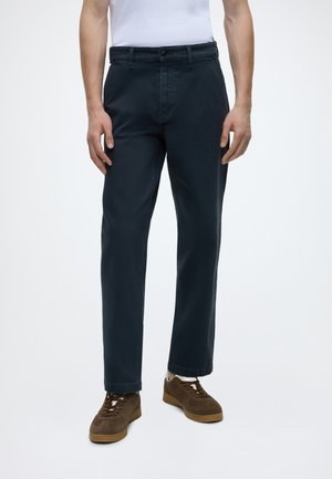 Mann trägt dunkelblaue gerade geschnittene Hose, weißes Hemd und braune Schnürsneaker und steht vor einem schlichten hellen Hintergrund.