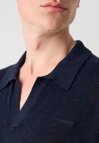 Marineblå teksturert polo skjorte med krage og GANT-logo på venstre side. Nærbilde med fokus på halslinje og stoffdetaljer.