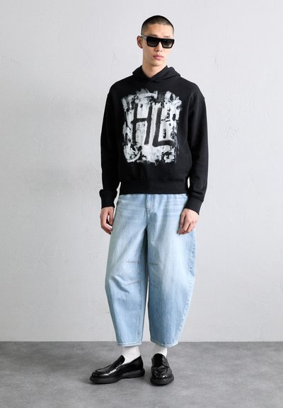 Sudadera negra con diseño gráfico blanco, vaqueros holgados azul claro con costuras laterales y zapatos negros sin cordones. El modelo lleva gafas de sol.