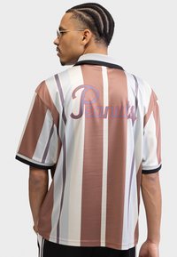 Short-sleeve shirt featuring vertical stripes in brown, cream, and gray. Back displays the word "Peanuts" in red cursive. Black collar and cuffs.