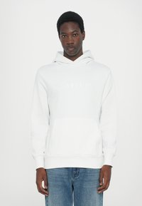 White hoodie with kangaroo pocket and elastic cuffs. Features embossed "QUARTZ CO" logo on chest. Matched with light blue denim jeans.