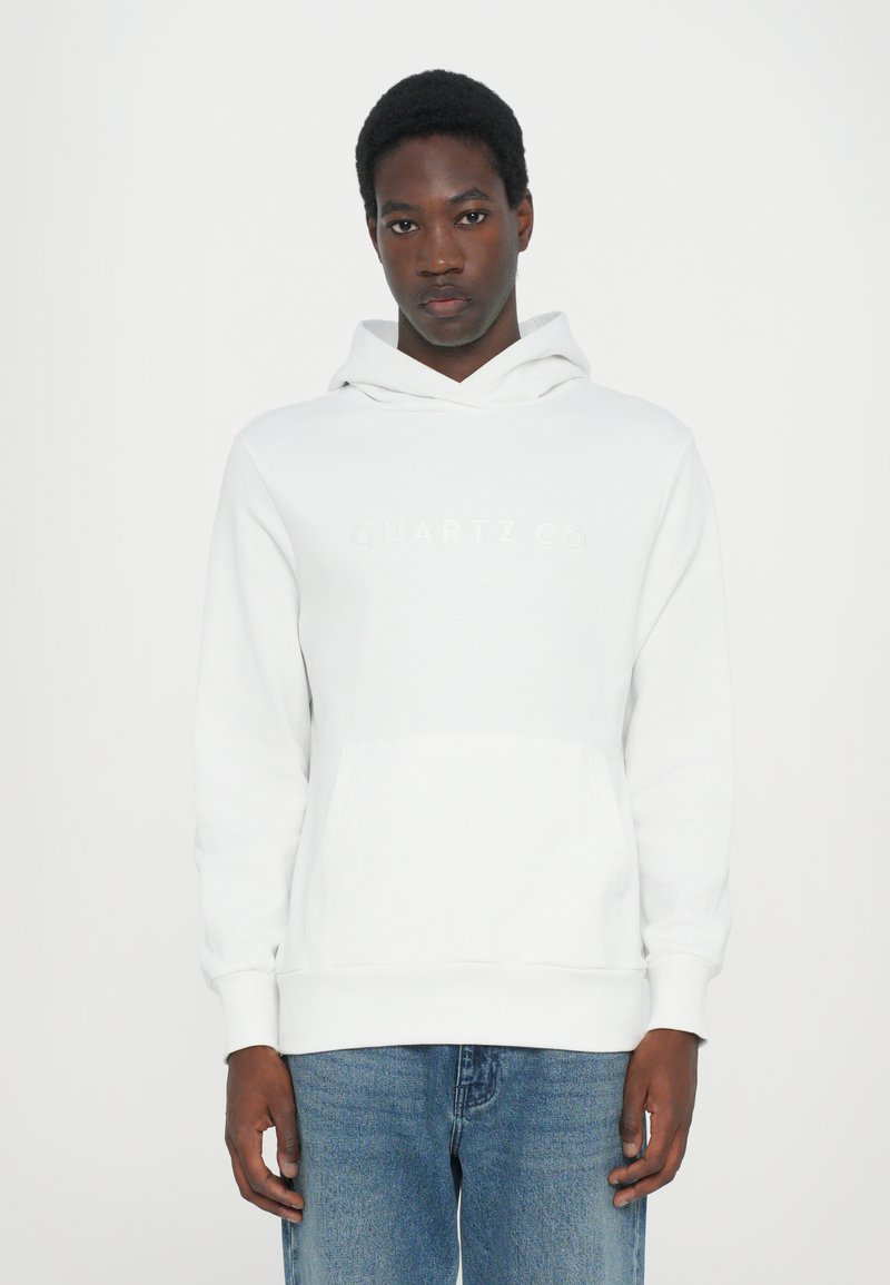 White hoodie with kangaroo pocket and elastic cuffs. Features embossed "QUARTZ CO" logo on chest. Matched with light blue denim jeans.