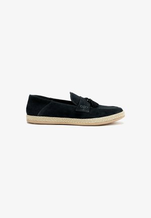 Black suede slip-on shoe with a flat woven jute sole and a decorative tassel on the front, featuring a rounded toe and minimal stitching.