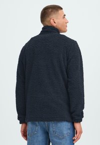 Granatowy polarowy sweter z wysokim kołnierzem i teksturowaną powierzchnią. Luźny fason, długie rękawy i brak widocznych zapięć z tyłu.