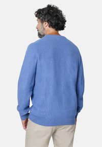 Hombre con cabello rizado que lleva un suéter texturizado de color azul claro y pantalones beige, de espaldas frente a un fondo gris claro.