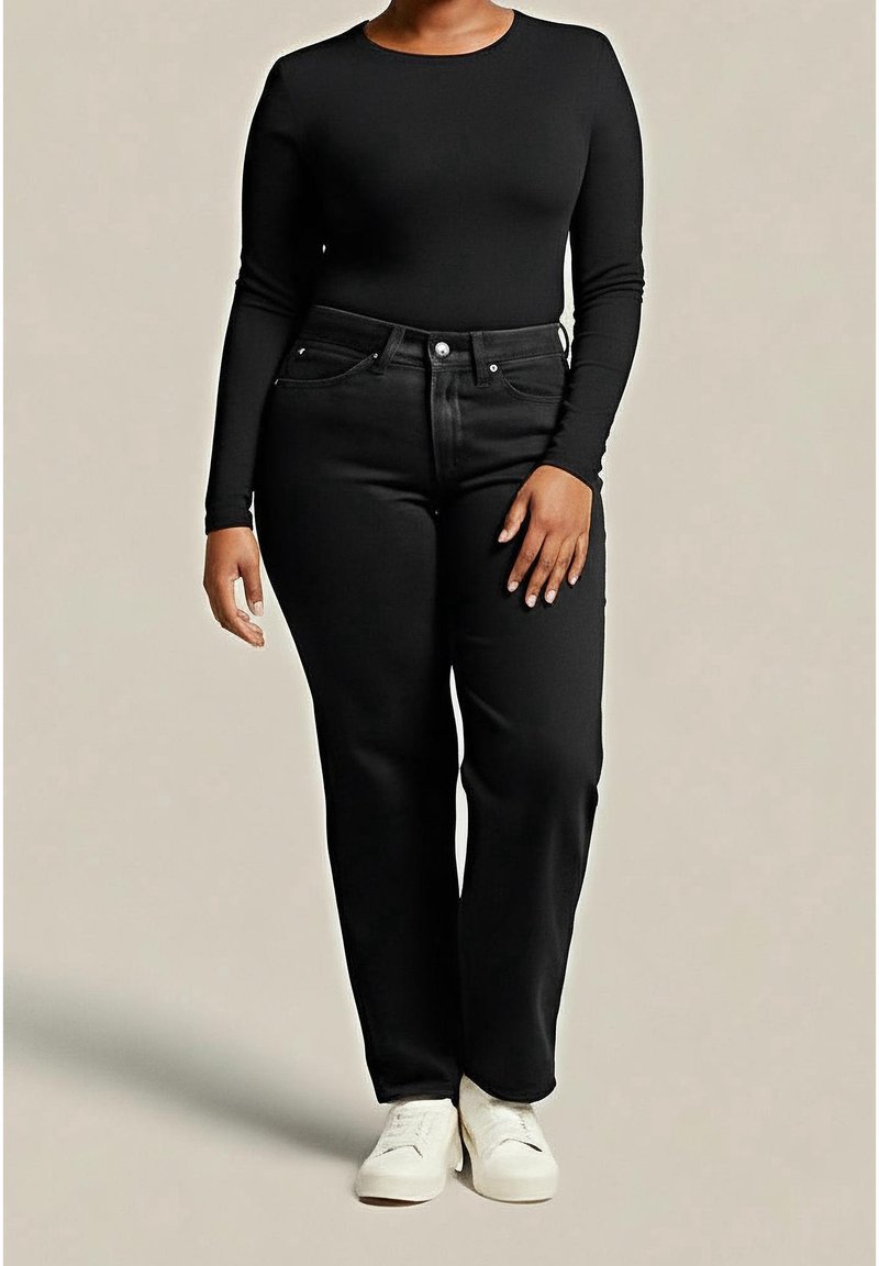 Personne portant un haut noir à manches longues, un jean noir taille haute et des baskets blanches, debout devant un fond beige uni.