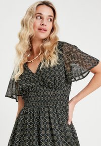 Schwarzes Kleid mit floralem Muster, kurzen transparenten Ärmeln, V-Ausschnitt, gerafftem Taillenbereich und fließendem Rock. Es weist komplexe grüne, rote und weiße Designs auf.