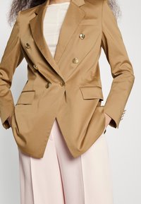 Femme portant un blazer croisé beige avec des boutons dorés, un top blanc et un pantalon large rose clair sur un fond uni.