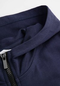 Granatowa bluza z kapturem na zamek błyskawiczny, o miękkiej teksturze, z wyraźnie zarysowanym kołnierzem i przednim zamkiem z czarnym uchwytem. Widoczne przeszycia.