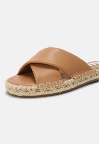 Sandale beige avec de larges brides en cuir entrecroisées et une semelle en jute tressé sur fond blanc, inclinée pour montrer les détails avant et latéraux.