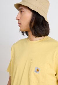 T-shirt en coton jaune avec une poche gauche sur la poitrine arborant un logo Carhartt carré. Un chapeau seau en coton beige clair complète l'ensemble.