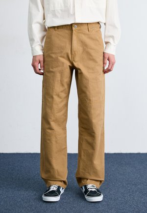 Pantalones de pierna ancha en color beige, confeccionados en una mezcla de algodón, con botón delantero, bolsillos laterales y una textura suave. Combinados con zapatillas negras.