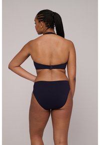 Vrouw met gevlochten haar in een donkerblauwe bikini, staande met één hand op haar heup, met de rug naar de camera tegen een effen lichte achtergrond.