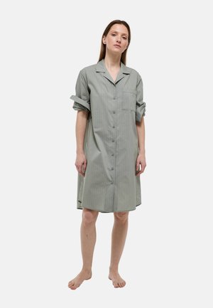 Femme debout, pieds nus, portant une robe grise ample jusqu'aux genoux, boutonnée, avec des manches retroussées et une poche poitrine, sur un fond blanc.