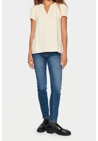 Krämfärgad kortärmad topp med V-ringning och strukturerat mönster, i kombination med åtsittande blåjeans och glänsande svarta loafers.