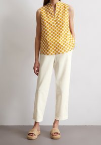 Top sin mangas amarillo con estampado y escote en V, pantalones rectos color crema y sandalias de lona beige, en una persona de pie.