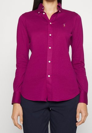 Chemise violette à boutons blancs avec un petit logo brodé sur le côté gauche de la poitrine, portée avec un pantalon foncé par une personne debout.