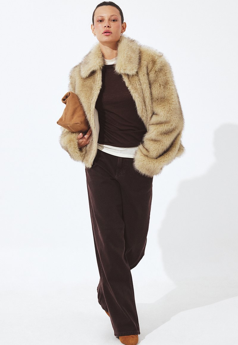 Falošná kožušina v béžovej farbe s širokým golierom, nosená cez tmavohnedý top a hnedé kordové nohavice; drží béžový clutch. Svetlé pozadie.