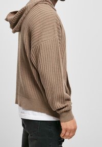 Hoodie en maille côtelée marron avec manches longues, coupe décontractée et surface texturée. Porté sur une chemise blanche, il comporte une capuche et des coutures latérales.