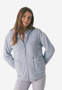 Helles, flauschiges, zip-fähiges Kapuzenoberteil in Blau mit zwei Vorder taschen und einem kleinen Logo. Trägt man über einem karierten Schlafanzugshirt, das eine weiche Textur aufweist.