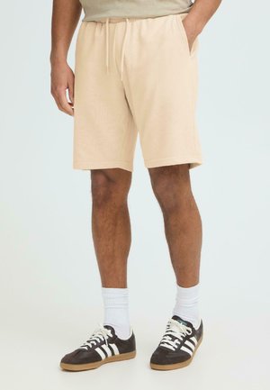 Mężczyzna w beżowych szortach z sznurkiem, białych skarpetkach do kostek i czarnych butach Adidas z białymi paskami na jasnym, gładkim tle.