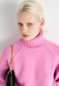 Pink knitted turtleneck sweater with ribbed details and a chunky texture, paired with a black handbag featuring a gold chain strap.