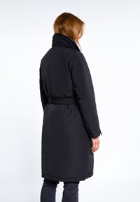 Vrouw met rood haar in een met een riem afgewerkte zwarte lange winterjas, staande in profiel tegen een effen witte achtergrond.
