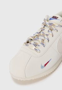 Nike kedas krēmkrāsas ādā ar daudzkrāsainiem auda diegiem. Sānos ir zils un sarkans izšūts logotips un teksturēta zole.