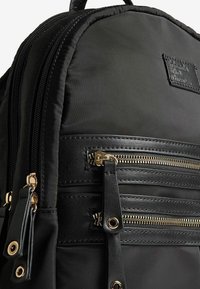 Sac à dos en nylon noir avec des garnitures dorées, deux poches zippées à l'avant et un accent en cuir texturé. Forme arrondie avec des bretelles ajustables.