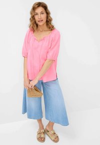 Top di lino rosa con maniche a puff corte e scollatura a V, abbinato a culotte di denim blu chiaro e sandali multicolori. Tiene una borsa a tracolla intrecciata.