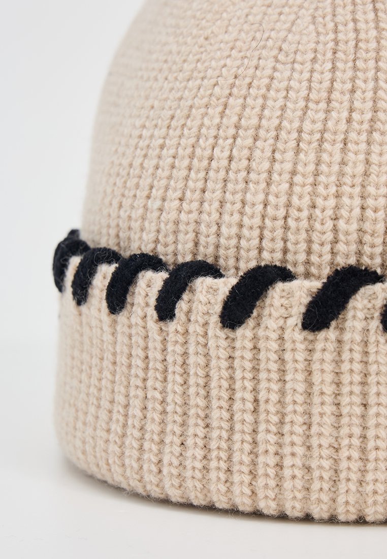 Beige Strickmütze mit gerippter Textur und einem schwarzen wellenförmigen Akzentband am Rand. Der Stoff wirkt weich und warm.