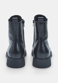 Un paio di stivali neri in pelle alla caviglia con zip laterali e linguette di tiraggio, visti da dietro su uno sfondo bianco.