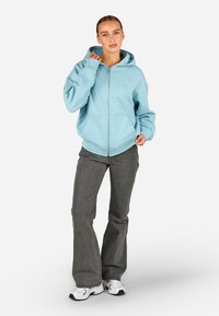 Világoskék cipzáras kapucnis pulóver, puha anyagból készült, kapucnival, kenguruzsebekkel és bordás mandzsettával. Szürke trapéz fazonú farmernadrággal és fehér sneakerrel párosítva.