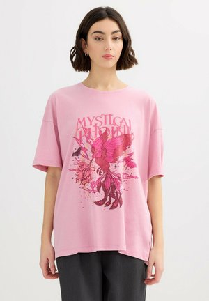 Mujer con una camiseta rosa holgada con un gráfico detallado de un fénix rosa y el texto "Fénix Místico", combinada con pantalones oscuros.