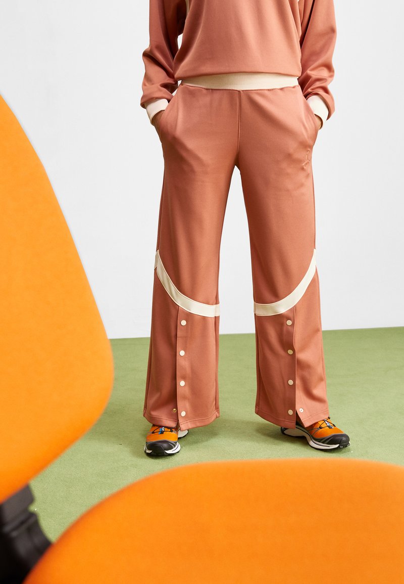 Pantalons de sport larges couleur pêche clair avec des accents blancs, des boutons pression et des poches latérales, associés à des chaussures de sport orange et noires. Fond en moquette verte.