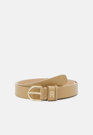 Ceinture en cuir tan enroulée avec une boucle en métal doré et un petit emblème logo doré sur la passants de ceinture sur fond blanc.
