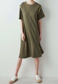 Olive green midi dress in soft fabric with short sleeves, ribbed neckline, and yellow accent stitching near shoulders. Paired with white sneakers.