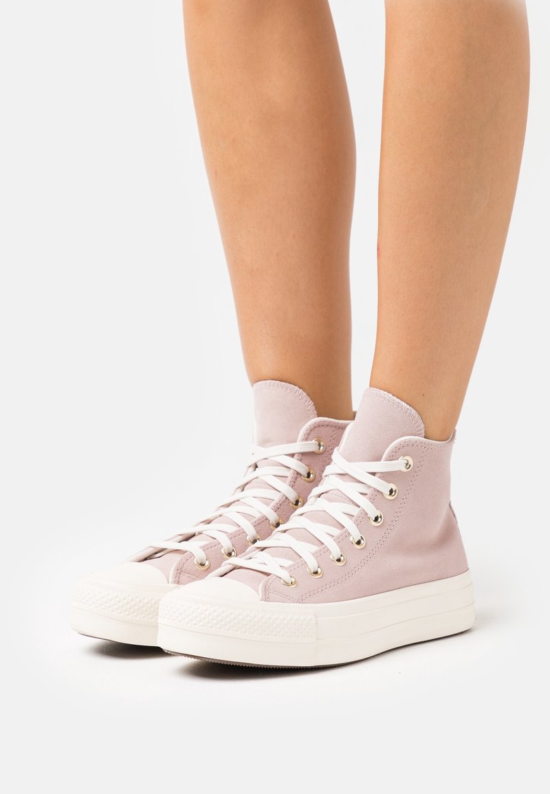 Jambes portant des baskets montantes rose clair avec des lacets blancs et des semelles épaisses en caoutchouc blanc sur fond blanc.