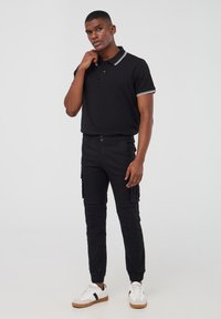 Czarna koszulka polo z białym paskiem przy kołnierzyku, połączona z czarnymi spodniami cargo i białymi trampkami z ciemnymi akcentami. Model stoi w swobodnej pozycji.