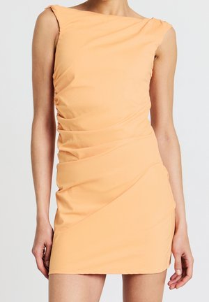 Mouwloze perzikkleurige geplooide mini-jurk met een asymmetrische halslijn gedragen door een staand persoon tegen een effen achtergrond.