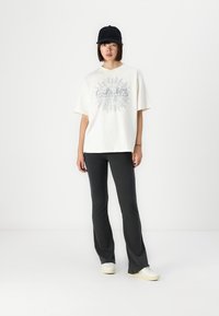 Elfenben oversized t-shirt med sølv grafisk design, sort baseball cap og mørkegrå flared bukser, parret med hvide lavt top sneakers.