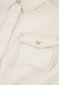 Weiße Denim-Hemd mit spitzem Kragen, einer einzelnen Brusttasche mit einem herzförmigen Knopf und kontrastierenden Nähten.