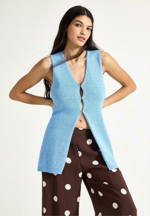 Giovane donna che indossa un gilet azzurro a maglia senza maniche con scollo a V profondo, abbinato a pantaloni marroni a gamba larga con grandi pois bianchi.